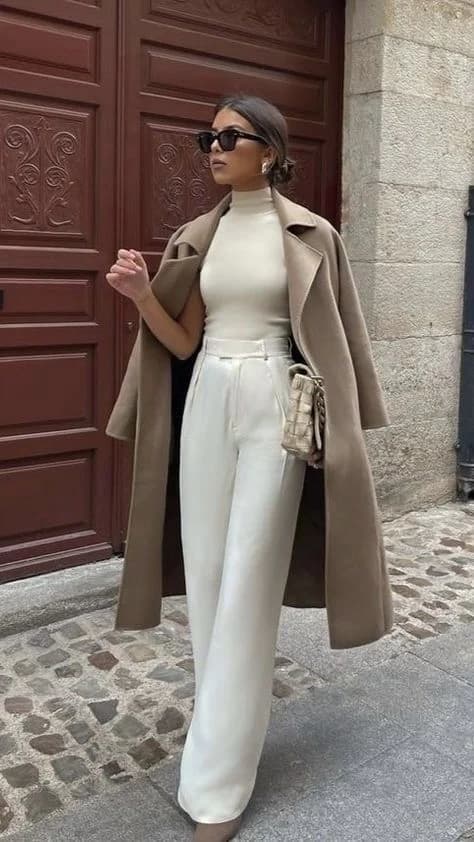 Mujer con abrigo camel sobre los hombros, top crema y pantalón blanco de pierna ancha frente a una puerta de madera